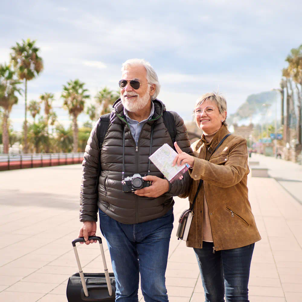 Uśmiechnięta para seniorów z walizką i aparatem spaceruje po deptaku w słonecznym kurorcie, przedstawiając spokojne wyjazdy i zwiedzanie dla osób starszych z Travel Well